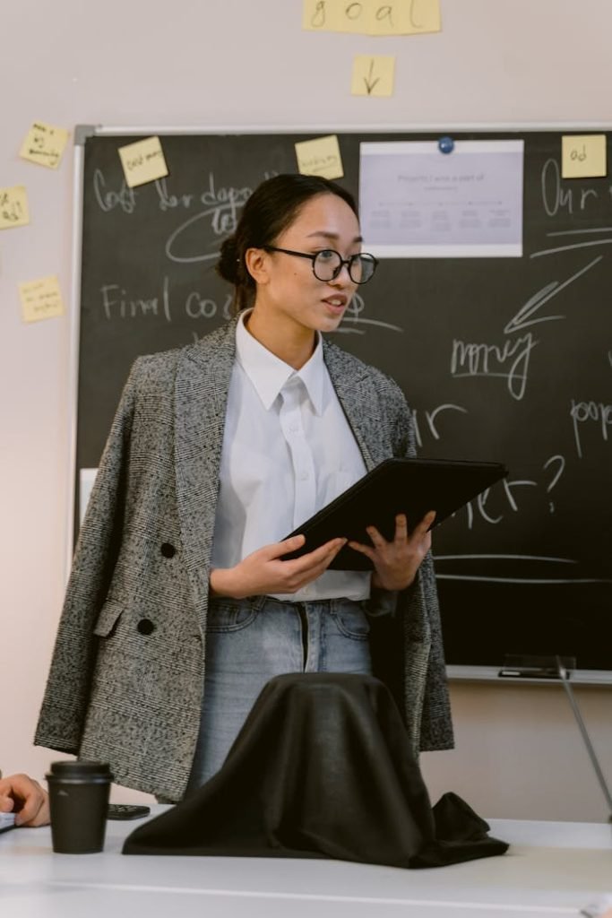 Asian woman presenting in an office setting in front of a chalkboard with notes.