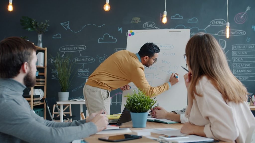 Arab man is writing on whiteboard and speaking to colleagues in creative shared office during briefing on marketing plan. People, business and teamwork concept.